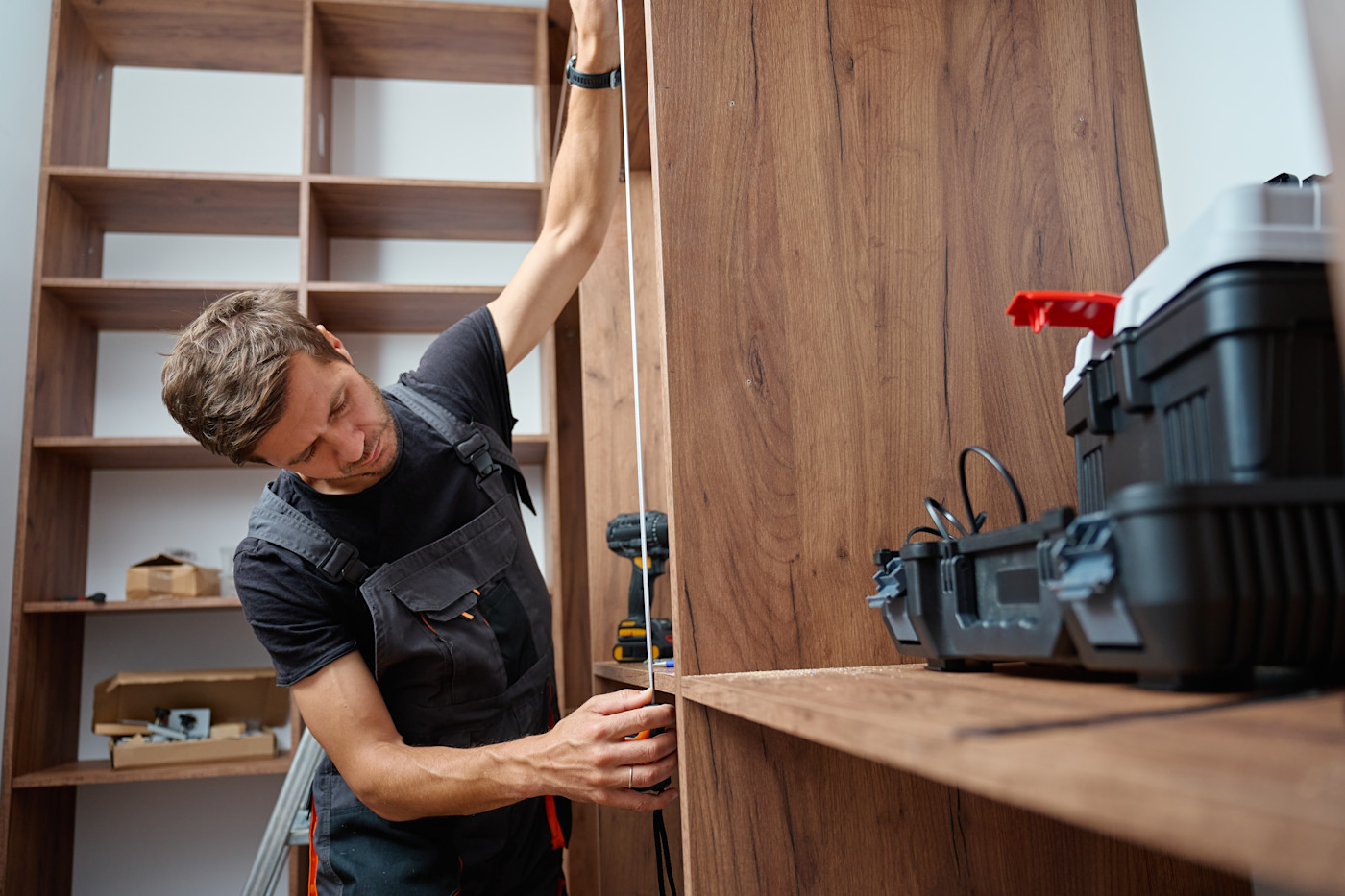 This image shows a carpenter using a measuring tape while fitting mid-tone wood wardrobes in a room. He is wearing overalls and there is a tool kit in the frame.