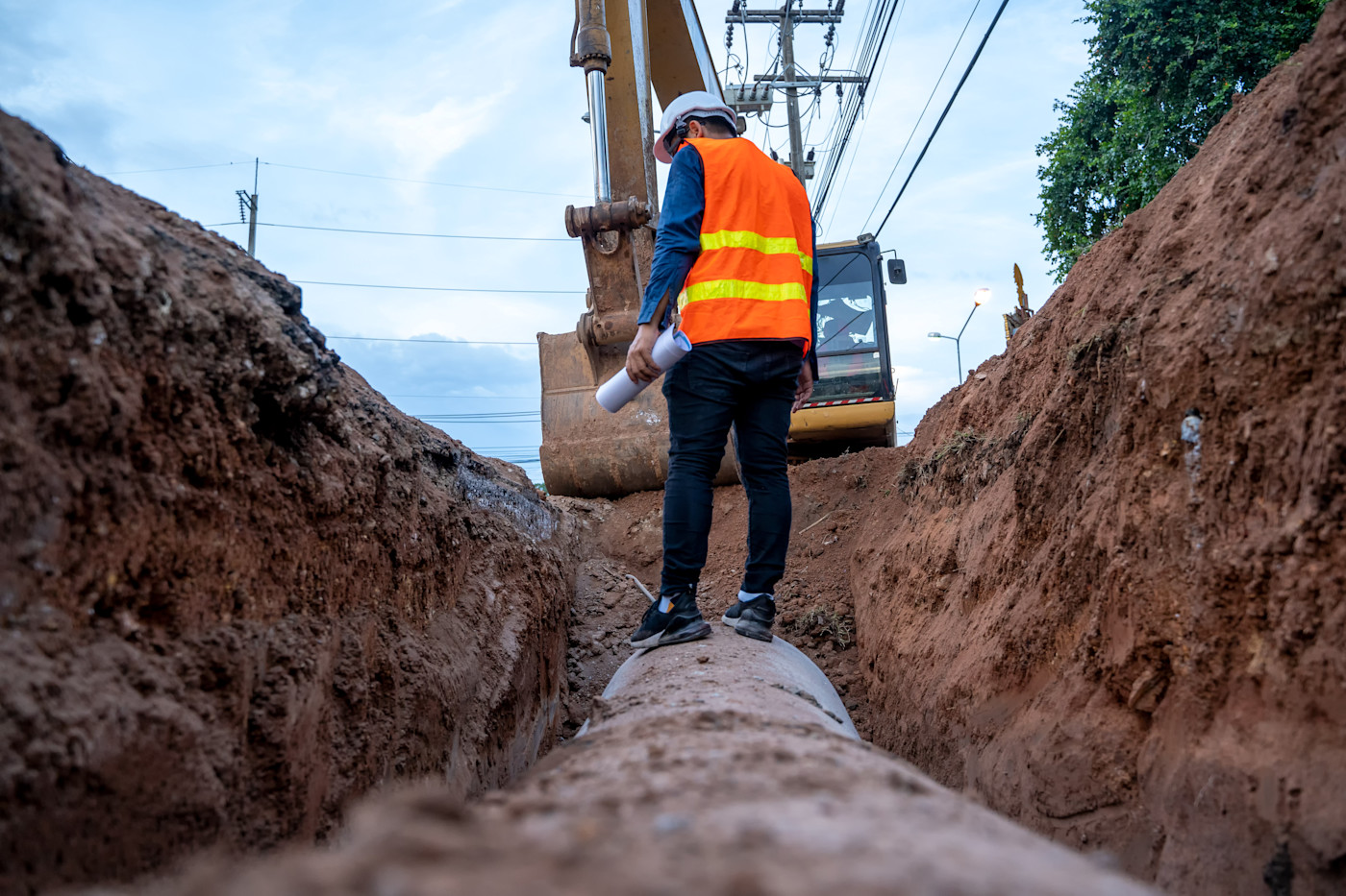This image shows a groundworker wearing a high vis jacket and hard hat standing atop of a pipe in an excavated trench