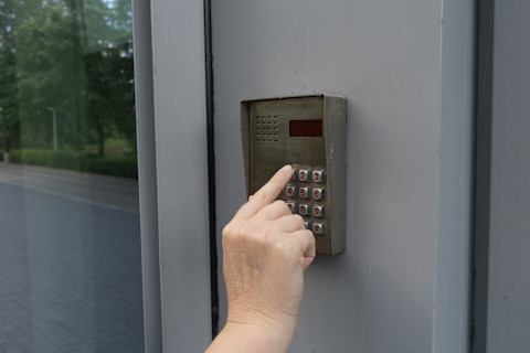 A close-up of a person’s hand entering a code on a metal keypad access control system mounted on a gray wall or door. The keypad includes numbers 0-9, a star (*), and a pound (#) sign. The hand is pressing the number 2. The environment outside reflects on the glass surface adjacent to the keypad, revealing a tree-lined street with a sidewalk, indicating that the entry system might be for a secured building or gated area. The overall scene suggests a focus on security and controlled access.