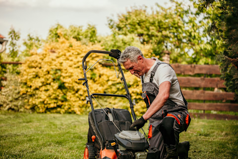A middle-aged man is kneeling on the grass in a backyard while working on a lawn mower. He is wearing work overalls and gloves, indicating that he is performing maintenance or repair on the machine. The setting is outdoors with a garden in the background, featuring green trees and yellow bushes. The man appears focused on his task, and the image conveys a sense of hands-on repair work.