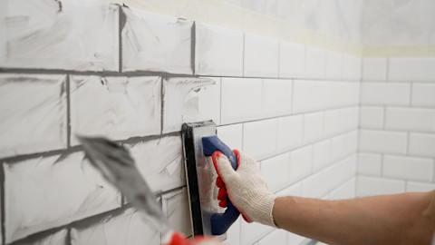 A person wearing gloves applies grout to white subway tiles on a wall using a float, precise and focus. Dark grey grout is spread visibly across the white tiles.