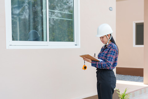 Young building inspector in hard hat taking notes on clipboard while inspecting building exterior.