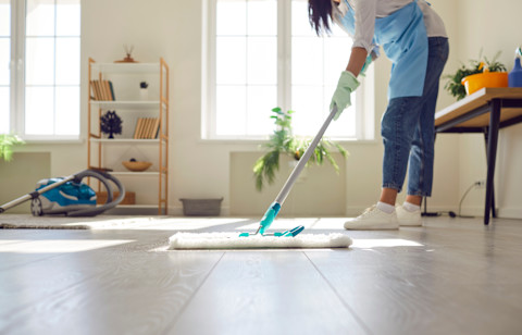 This image shows a professional cleaner wearing an apron and rubber gloves mopping a hardwood floor. There is a vacuum cleaner in the background.