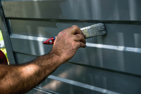 Close up of the forearm and hand of a man as he adds a new layer of grey paint to an exterior garage door with a small paintbrush.