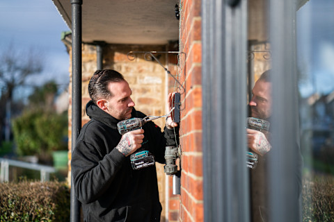 An electrician standing in a porch of a building with a drill in hand fixing an outdoor socket.