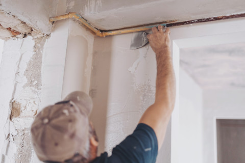A male plasterer wearing a baseball cap reaches up to the top of a wall of a house under renovation to spread plaster using a finishing tool.
