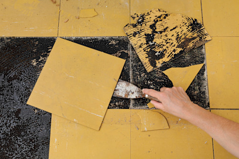 A hand uses a scraper to remove damaged yellow linoleum tiles from a floor during a renovation project, revealing blackened adhesive underneath.