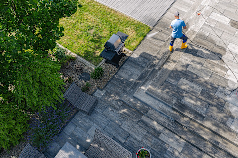 A person uses pressure washer to clean and maintain a stone patio. Grass and garden are visible nearby under clear sky.