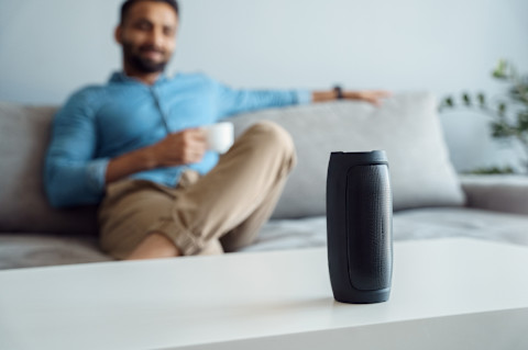 Close-up of a man sitting and listening to a smart speaker. The man is relaxed, with the smart speaker placed on a nearby table. The image shows the speaker in use, with the man enjoying audio playback or interacting with the device.