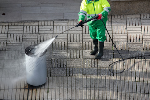 A professional bin washing service in action, showing a worker cleaning a large outdoor waste bin using a high-pressure washing machine. The setup includes a specialized vehicle equipped with cleaning tools, highlighting the bin washing process and the professional equipment used to maintain hygiene.