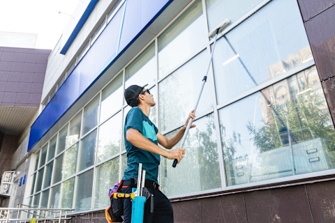A window cleaner using a squeegee and cleaning solution on a large glass window, with a clear view of the outdoors and a ladder positioned nearby.