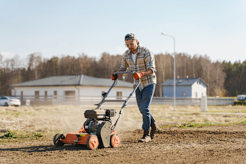 Close-up of a lawn aeration service in progress. A worker is using a core aerator machine to punch holes into a grassy lawn, with soil plugs visible on the surface. The background shows a well-maintained yard with green grass and trees