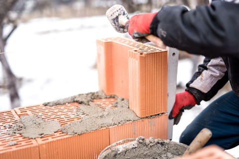 This image shows a mason or bricklayer working on a snowy construction site, carefully placing bricks and applying mortar to build a sturdy wall.