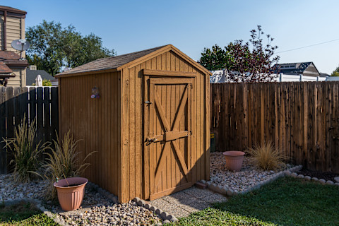 A large wooden shed in a back garden on gravel. Potted plants are beside the shed, and it is in front of a dark brown wooden fence.