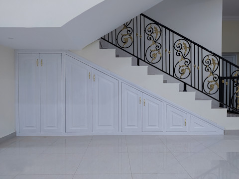 A neatly designed under-stairs storage unit with white cabinets, each door varying in size to fit the sloping space beneath the staircase. The cabinet doors are fitted with gold-colored handles. Above the storage unit, a staircase with decorative black wrought iron railings, embellished with gold accents, leads upwards. The area is clean and minimalistic, with a polished, light-colored floor that reflects the simple and elegant design of the space.
