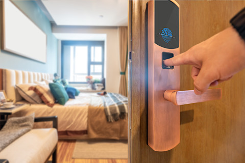 A close-up of a smart lock with a fingerprint scanner being used to unlock a door. A person's finger is placed on the scanner, and the lock features a modern design with a touchscreen interface. In the background, a stylish, well-lit bedroom with a neatly made bed and decorative pillows can be seen, emphasizing the convenience and security of smart lock technology in a home setting.