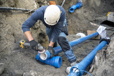 Close-up of a technician repairing a sewer line. The image shows the technician working in a trench, with exposed pipes and plumbing tools visible. The surrounding area includes earth and construction equipment.