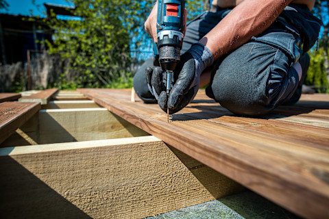 This image shows a deck installer using an electric screwdriver to install wooden decking boards during deck construction.