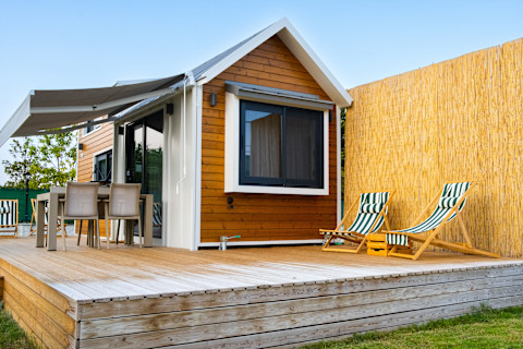 A small dark brown wooden cabin house on a raised brown deck against a fence in a garden. Two sun loungers and a dining table on chairs are beside the house on the decking.