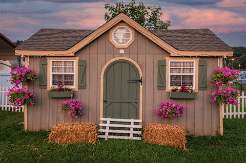 This image shows a garden shed with a shingle roof, windows, hanging baskets with pink flowers and hay bales by the door.