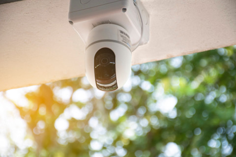 A security camera is mounted on the ceiling outdoors, aimed downward. The camera is white, sleek, and modern, likely equipped with advanced features such as motion detection or night vision. The background shows a natural outdoor setting with blurred green foliage and sunlight, creating a peaceful atmosphere while highlighting the importance of surveillance for security purposes.