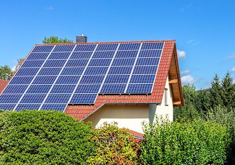 This image shows a number of solar panels on the roof of a house. The roof is made from red/orange tiles and it contrasts against the off-white exterior of the home. There are full, green bushes in the foreground obscuring the bottom half of the property. There is a blue sky and some white clouds.