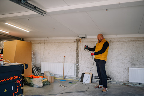 Man in yellow vest operates a power tool inside a garage, surrounded by cardboard boxes and various construction materials scattered on the floor