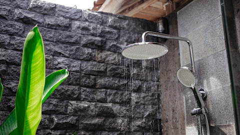 A tropical outdoor running shower head against the background of a grey brick wall with green plant leaves growing from the left.