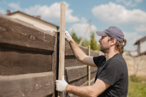 This image shows a tradesperson wearing safety gloves fixing a section of brown wooden fencing in a garden.