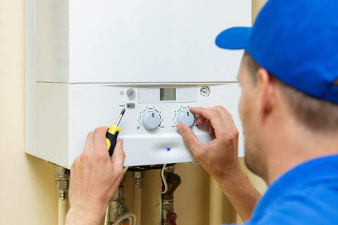 A technician in a blue cap is adjusting the dials on a white home heating boiler with a digital display, using a screwdriver on the reset button.