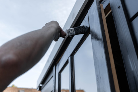 A man painting the exterior of a shed, above the door windows, using a midnight black colour