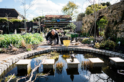 A man dressed in black is crouching on the ground in front of a pond, with a yellow bucket next to him. He is in a garden filled with many plants and trees, holding a long tape measure as he works on the pond.