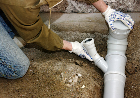 Close-up of a technician replacing a backwater valve. The image shows the technician working on the valve, with old and new valves visible. Surrounding pipes and tools are also visible, with a partially disassembled section of the plumbing system.