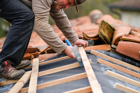A tattooed roofer is focused on installing or repairing a flat roof. He's in the process of laying or measuring a section of roofing material, possibly a metal or bitumen-based covering. The photo captures him in a crouched position, emphasizing the precision and attention to detail required for the task. The background reveals a partly cloudy sky and the silhouette of residential buildings, suggesting that the work is taking place in a suburban area. The roofer's clothing and visible tattoos on his arm add to the narrative of a skilled tradesperson at work.