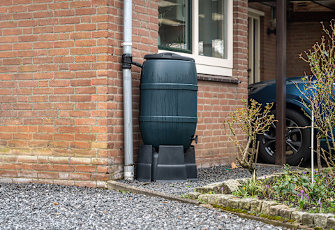 A large black rainwater harvesting container in the front garden of a domestic house. The container is lifted from a gravel pathway with a black, plastic stand, and pushed against the house wall beneath a window.