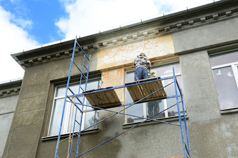 Worker standing on scaffolding in front of a building façade, applying or removing lime render on the exterior wall surface under a blue sky.