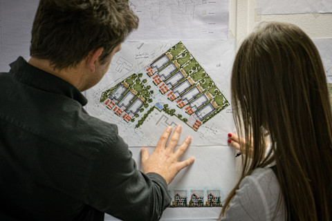 A man and a woman, presumably architects or planners, are attentively examining a colorful architectural site plan. Their fingers are placed on the paper, possibly pointing at specific details or areas of interest. The image shows the back of the man and over-the-shoulder view of the woman, both focused on the plan. The plan itself appears detailed with illustrations of housing units, landscaping, and layout annotations, indicating a residential development. The image captures a moment of professional collaboration and decision-making.