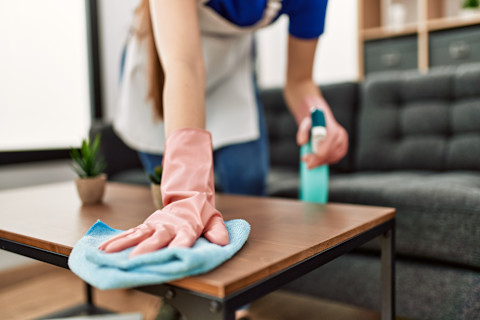This image shows a cleaning professional wearing rubber gloves to clean a coffee table with a cloth. She has a cleaning spray in her left hand.