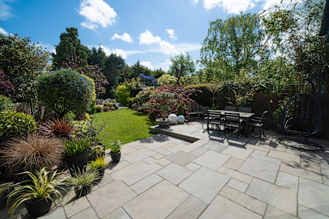 The image shows a lush green garden filled with plants, shrubs and trees and there is a paved patio. A wooden dining table sits on top of the patio, ready for alfresco meals.