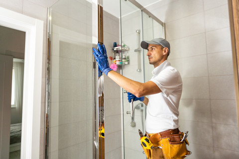 Close-up of a technician installing glass shower enclosures. The image shows the technician fitting glass panels into place with tools and mounting hardware visible. The background includes the shower area with partially installed enclosures.