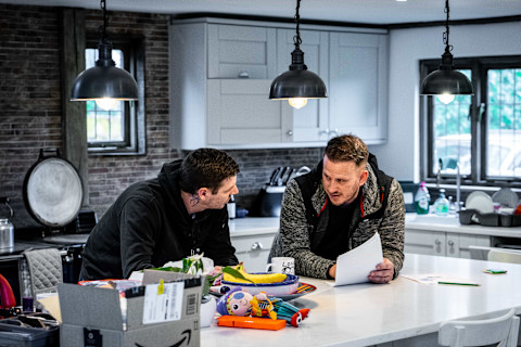 Two men are discussing something at a kitchen island. The man on the left, who has a tattoo on his neck, is leaning in attentively while the man on the right, holding a paper, is explaining something. The kitchen is modern, with hanging industrial-style pendant lights, grey cabinets, and various items scattered on the countertop, including a box and some children's toys.