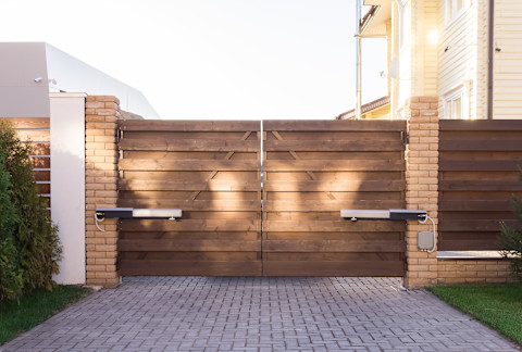 This image shows a wooden driveway gate at the end of a paved driveway. The gate is secured between two sand-coloured brick pillars. To the right and left of the image there is a glimpse of mowed green grass.