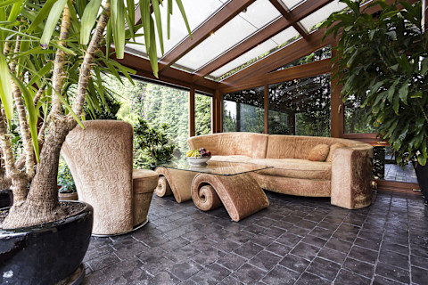 This image shows a stylish sunroom with dark tiled flooring and green potted plants. The window frames are made from a mid-tone wood and there is a light coloured sofa.