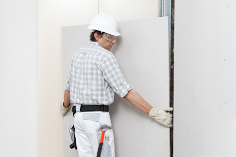 This image shows a tradesperson installing a plasterboard sheet on a wall in a partially finished room.