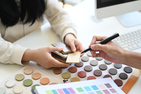 An interior designer giving advice to a client on colour samples at a desk, with one person holding material swatches and the other pointing with a pen near a palette of round paint colours.