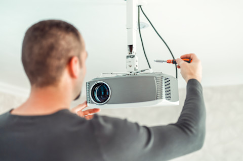 A man is in the process of setting up a projector in a living room, carefully connecting a cable to it.  The man is dressed in grey, and the living room has a clean, white backdrop, creating a bright and neutral environment that highlights the setup.