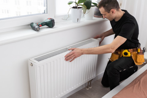 Plumber in a black uniform with a yellow utility belt with a screwdriver and a tape measure in the pockets works on a white domestic radiator situated under a window. On the window sill is a drill and three plants.