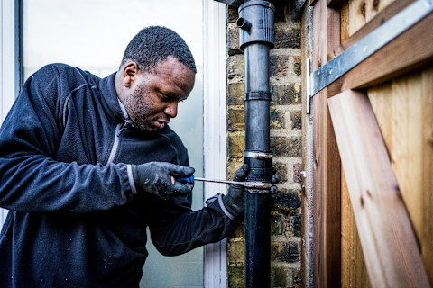 This image shows a plumber wearing protective gloves using a tool to fix an external leaking pipe on the side of a house.