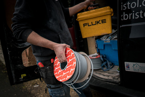 A close-up image shows a person in a work outfit holding a spool of electrical cable and a yellow tool case labeled 'FLUKE'. They are standing beside an open van filled with various tools and supplies. The person's tool belt is equipped with several screwdrivers, indicating they might be an electrician preparing or organizing equipment for a job. The van carries a decal suggesting it belongs to an electrical service company, as indicated by the NAPIT logo, a UK-based certification service for electrical technicians.