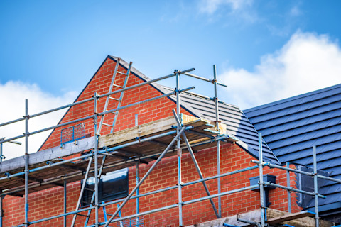 A red brick house with a black roof under construction, with scaffolding and a ladder against the front of the house. A cloudy blue sky is in the background.